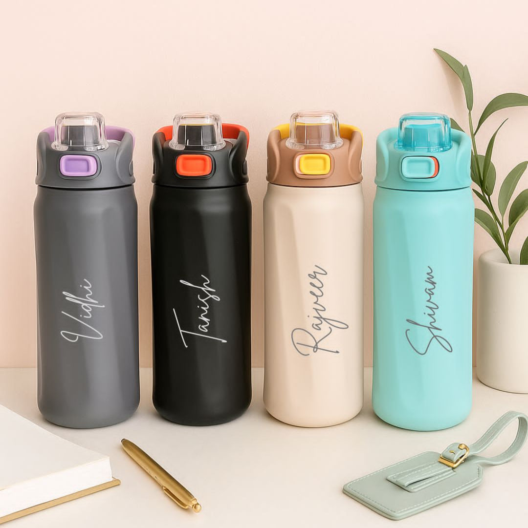 Four insulated water bottles in different colors with names engraved on them, placed on a surface with a plant in the background.